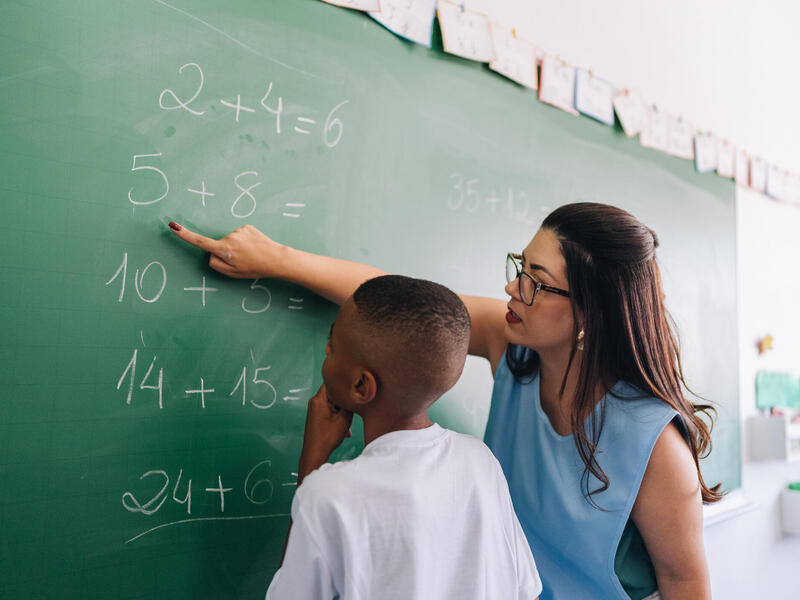 A photo of a woman pointing at a math problem on a chalk board. A boy is standing next to her looking at the problem. 