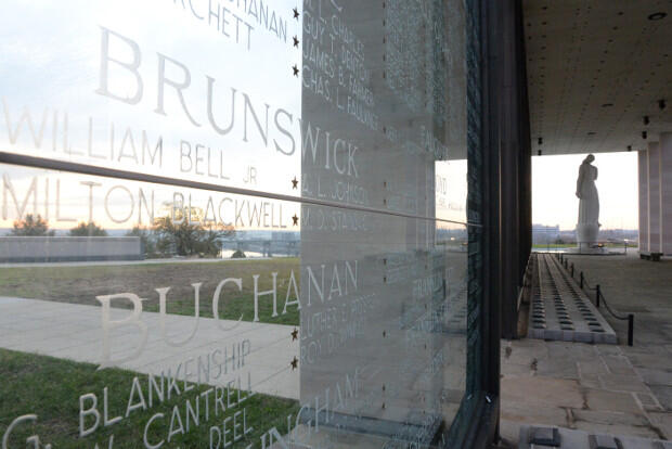 A view of a glass wall enscribed with names, with a statue in the background at right.