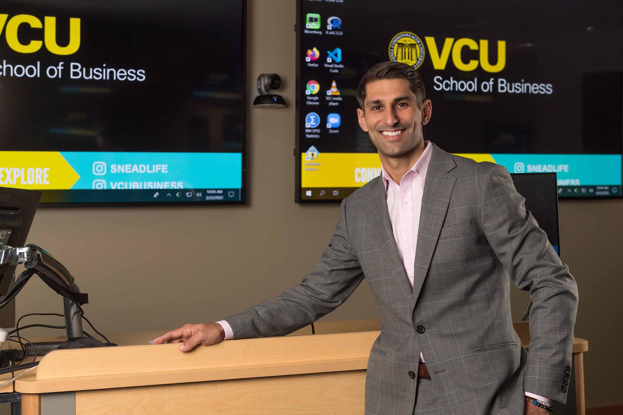 A photo of a man from the waist up. He is standing in front of a wooden podium and two large screens that read \"VCU\" in yellow letters and \"School of Business\" in white letters. 