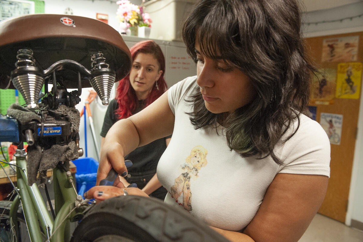 A woman working on a bike with another woman watching her. 
