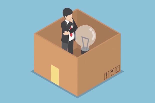 A figure stands inside of a cardboard box pondering a lightbulb.