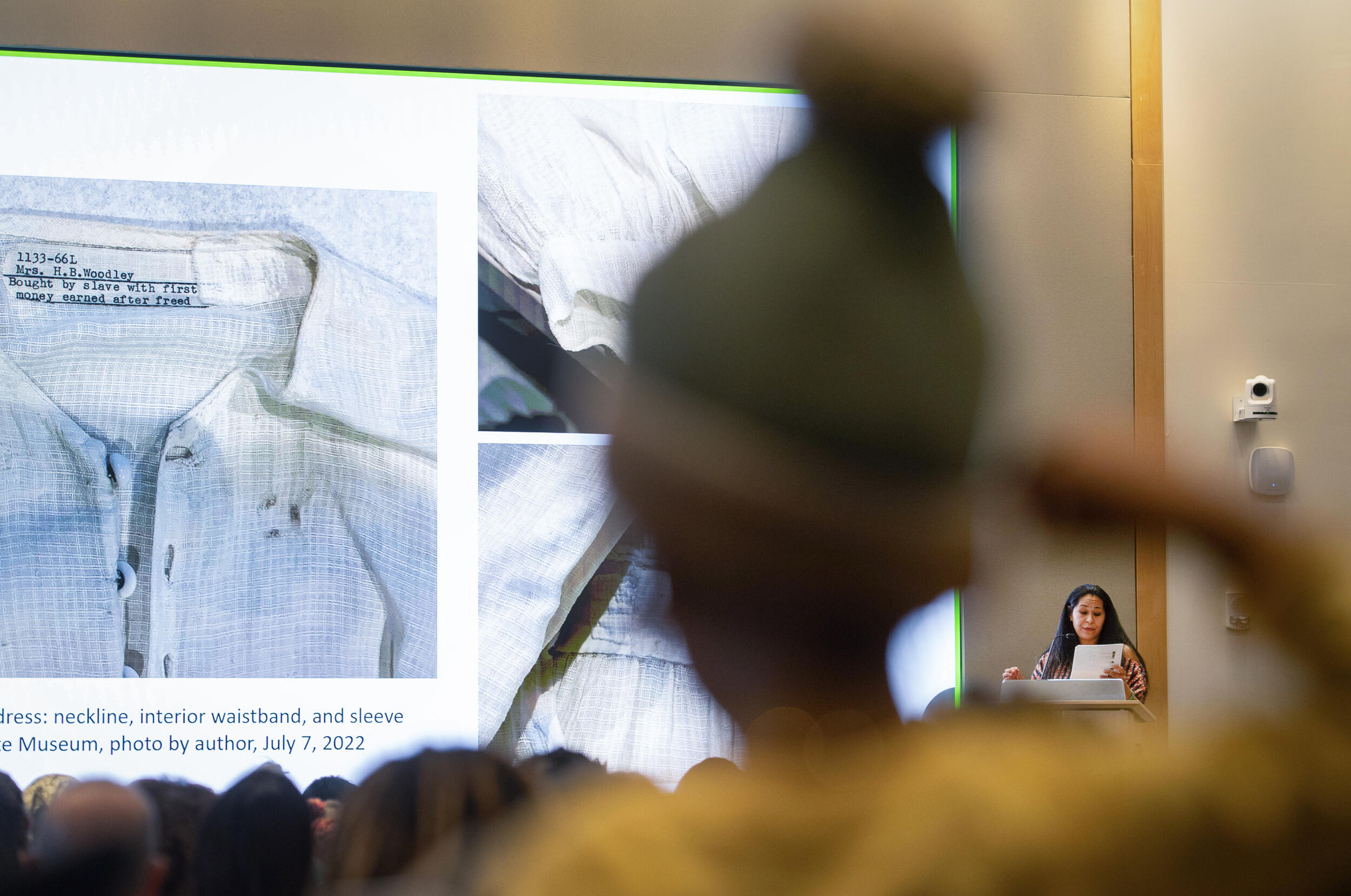 A photo of a crowd watching a woman speak at a podium. On the screen are close ups of a white dress. 