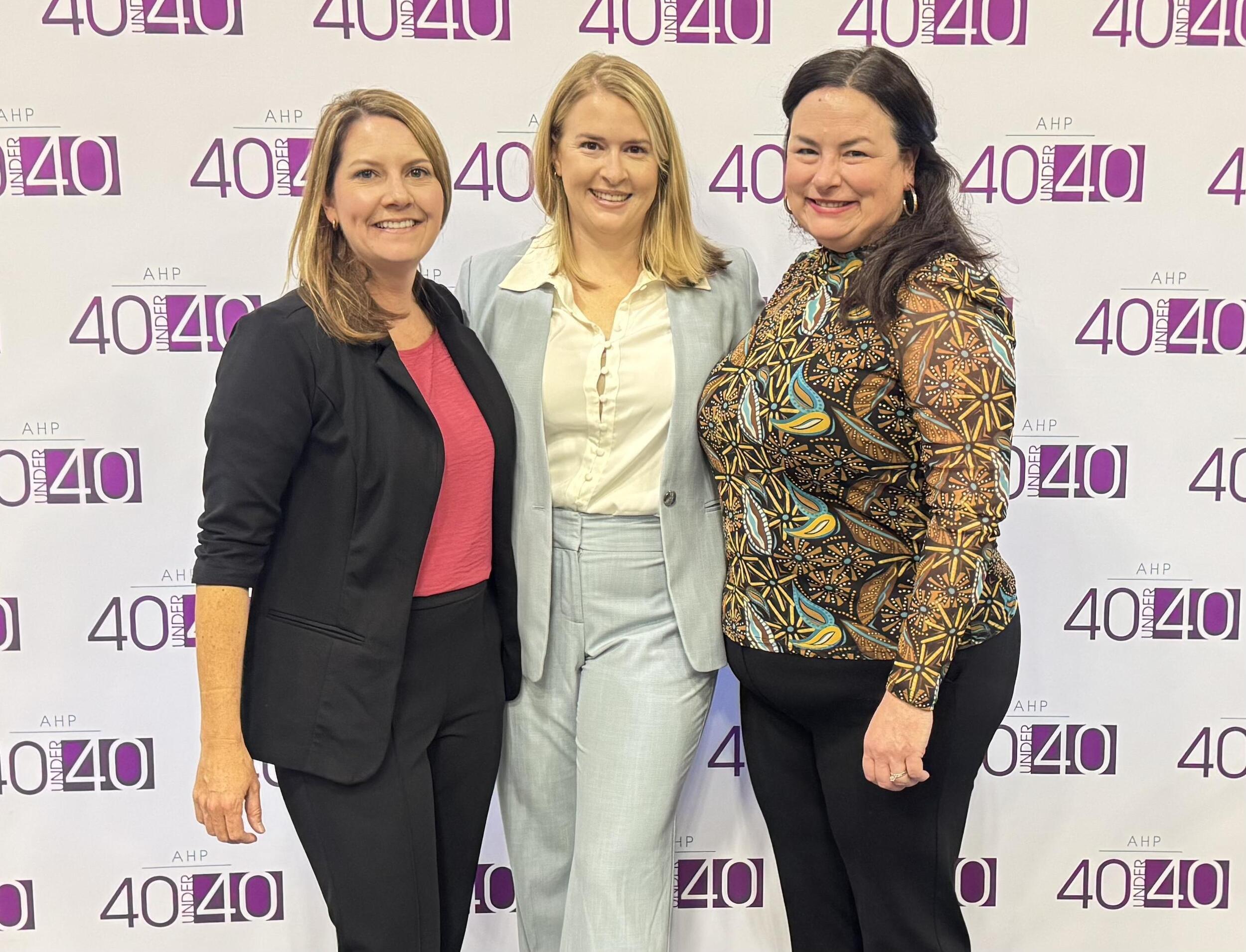 A photo of three women standing in front of a step and repeat that says \"40 40\" in purple print. 