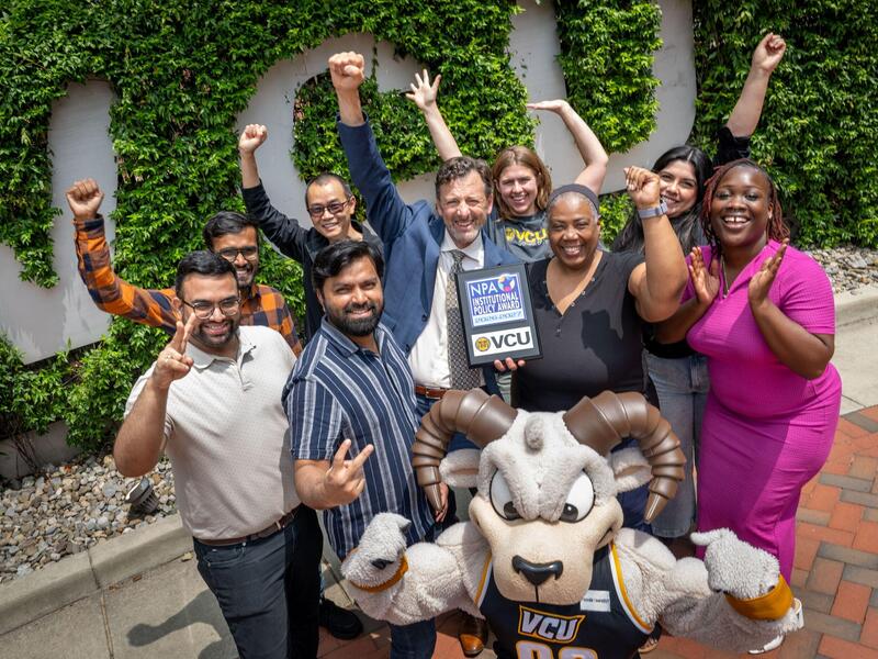 A group of people raised their arms in celebration with one man in the middle holding a plaque. A Ram mascot flexes in the front.
