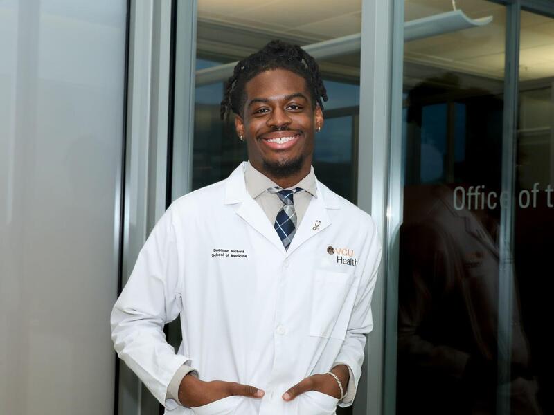 A photo of a man fromt he waist up. He is simling with his hands in his pockets and wearing a white lab coat. 