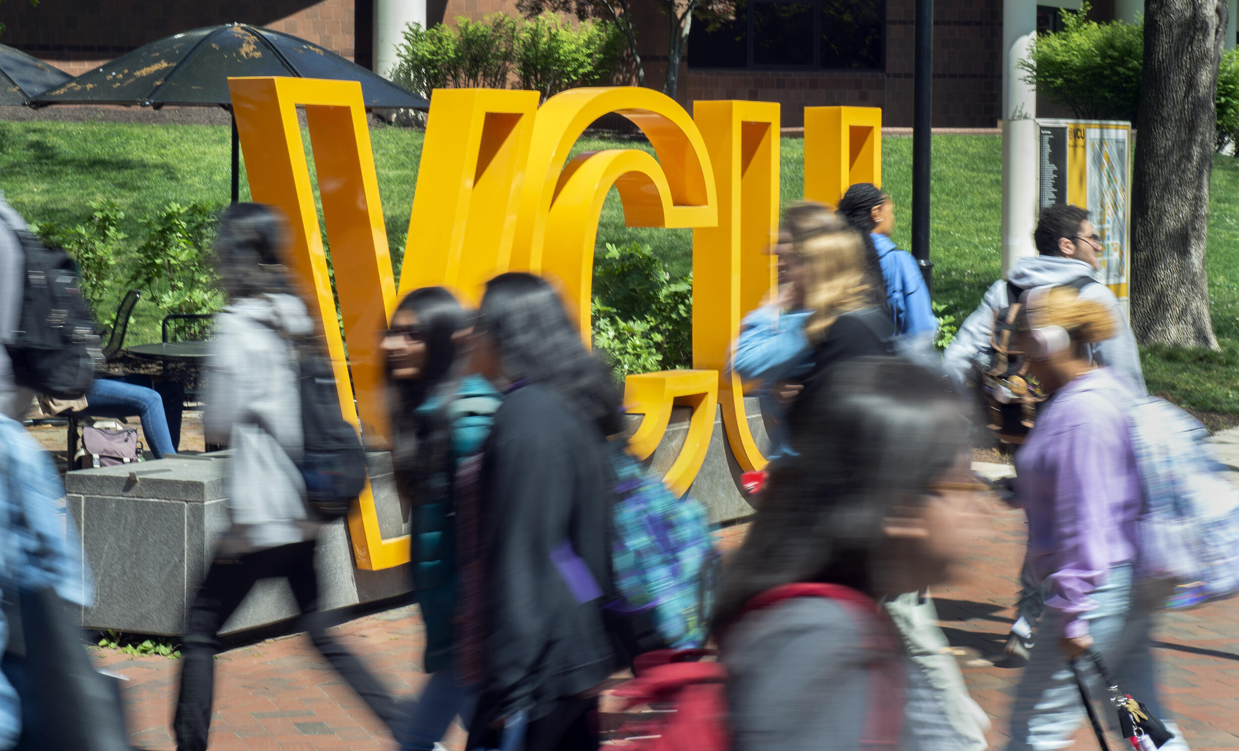 People walking across a sidewalk. There is a sign that spells out \"VCU\" in large yellow letters. 