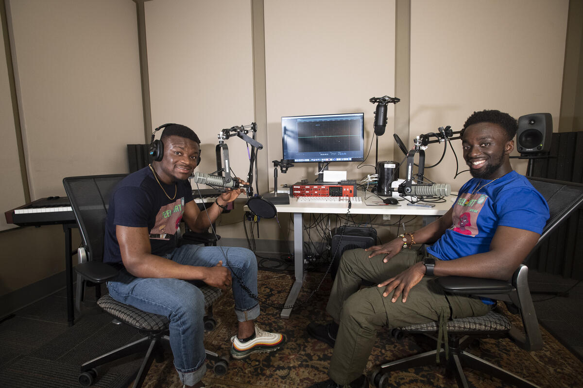 Roland Kusi and Atuahene Adu-Gyamfi sitting at a desk with podcasting equipment while wearing headphones