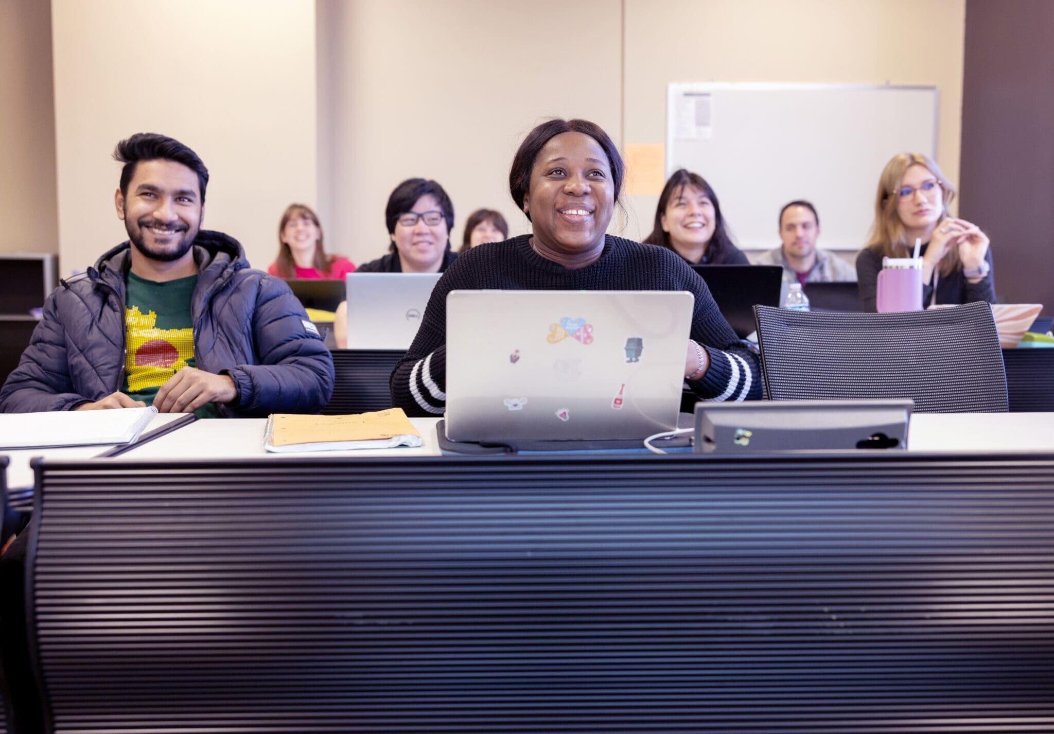 Students sit at long tables facing toward the front of a classroom space.