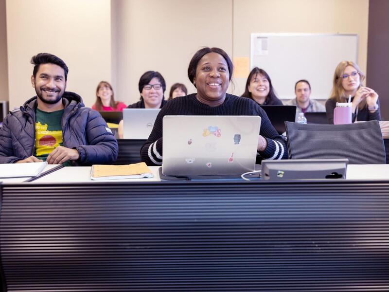 Students sit at long tables facing toward the front of a classroom space.