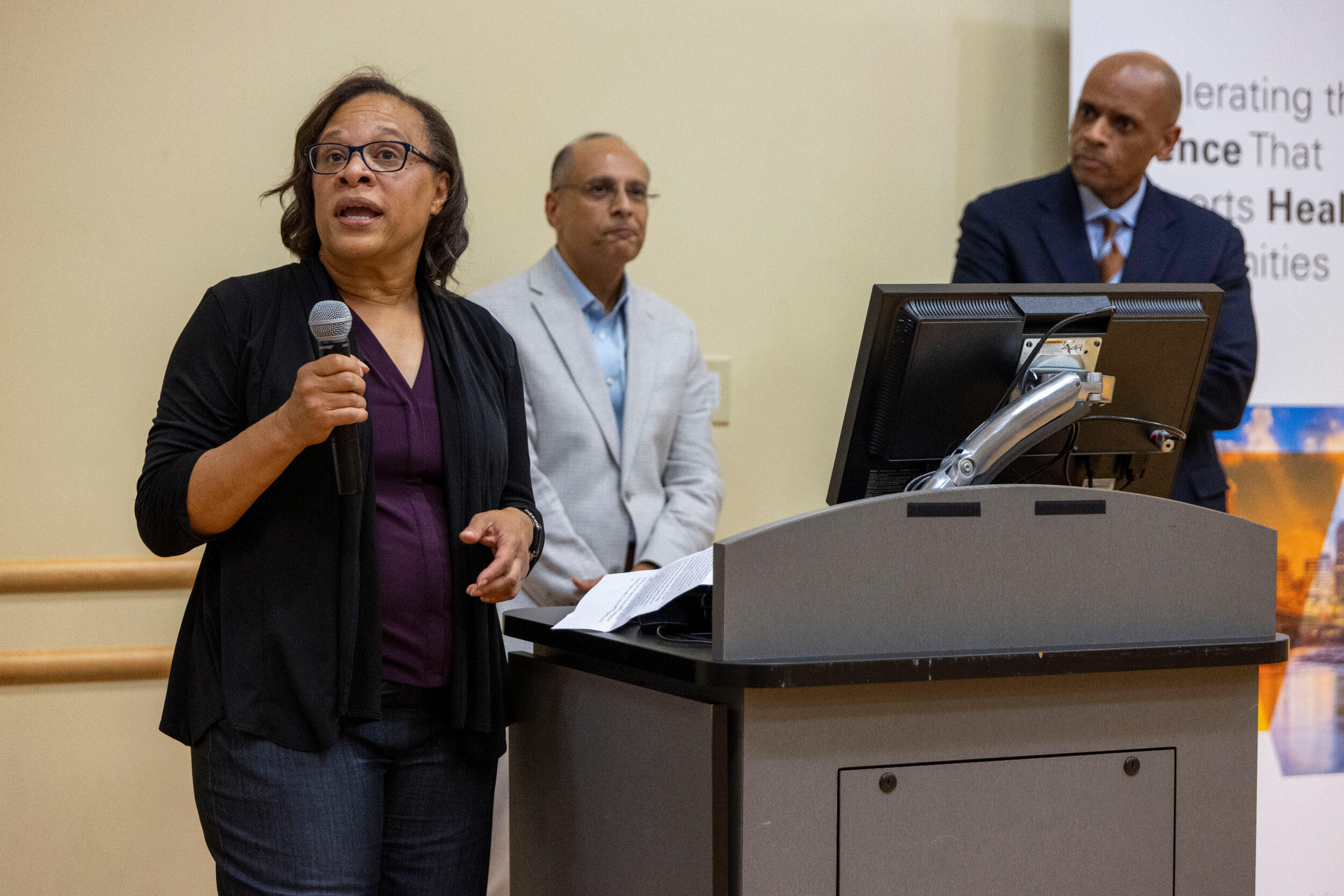 A woman speaks into a microphone while two men behind her listen.