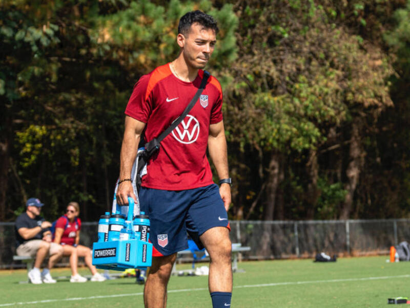 A man in a soccer uniform is standing on a field while holding a water bottle carrier and standing next to a ball. 