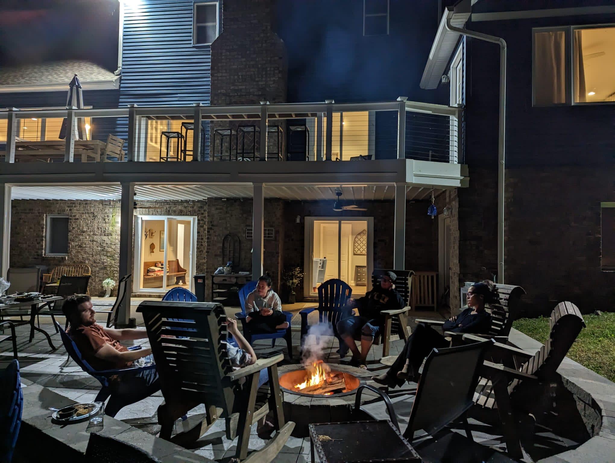 A photo of a fire pit with several chairs around it at nihgt. There are five people sitting around the fire. The fire pit is adjacent to a house. 