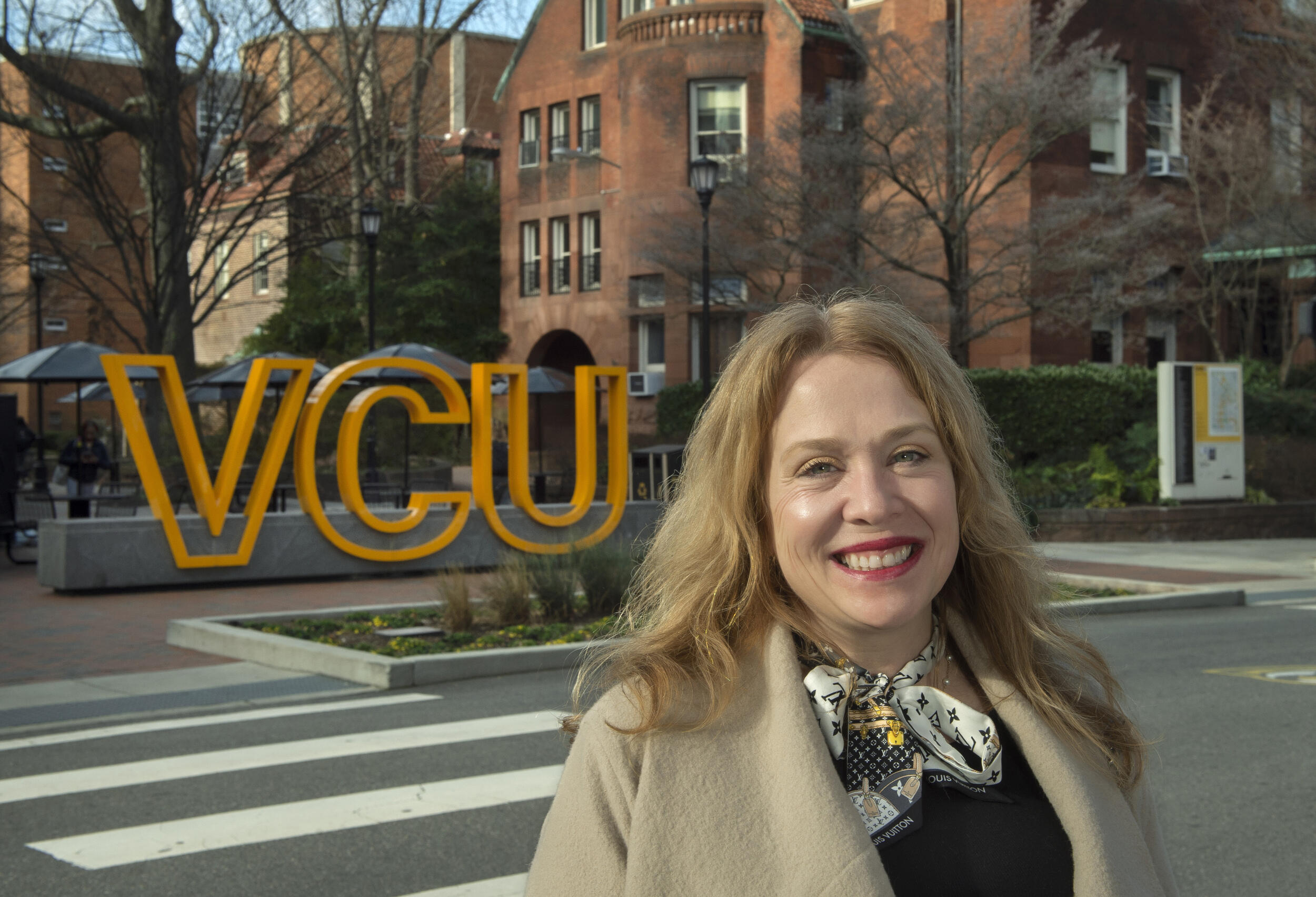 A photo of a woman from the shoulders up standing outside across the street from a sign that says \"VCU\" in yellow letters. 