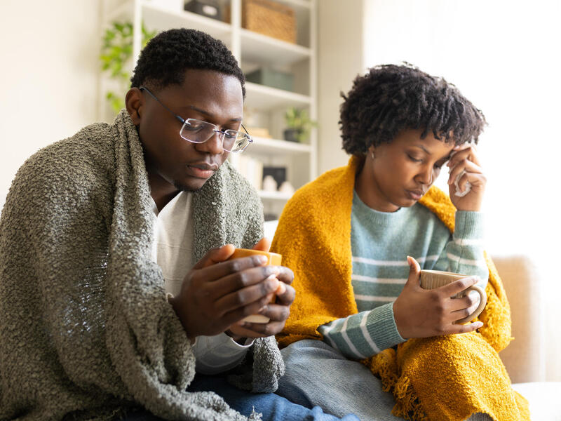 A man and a woman have blankets over their shoulders for warmth, appearing to feel ill, illustrating the pain experienced by sickle cell disease patients.