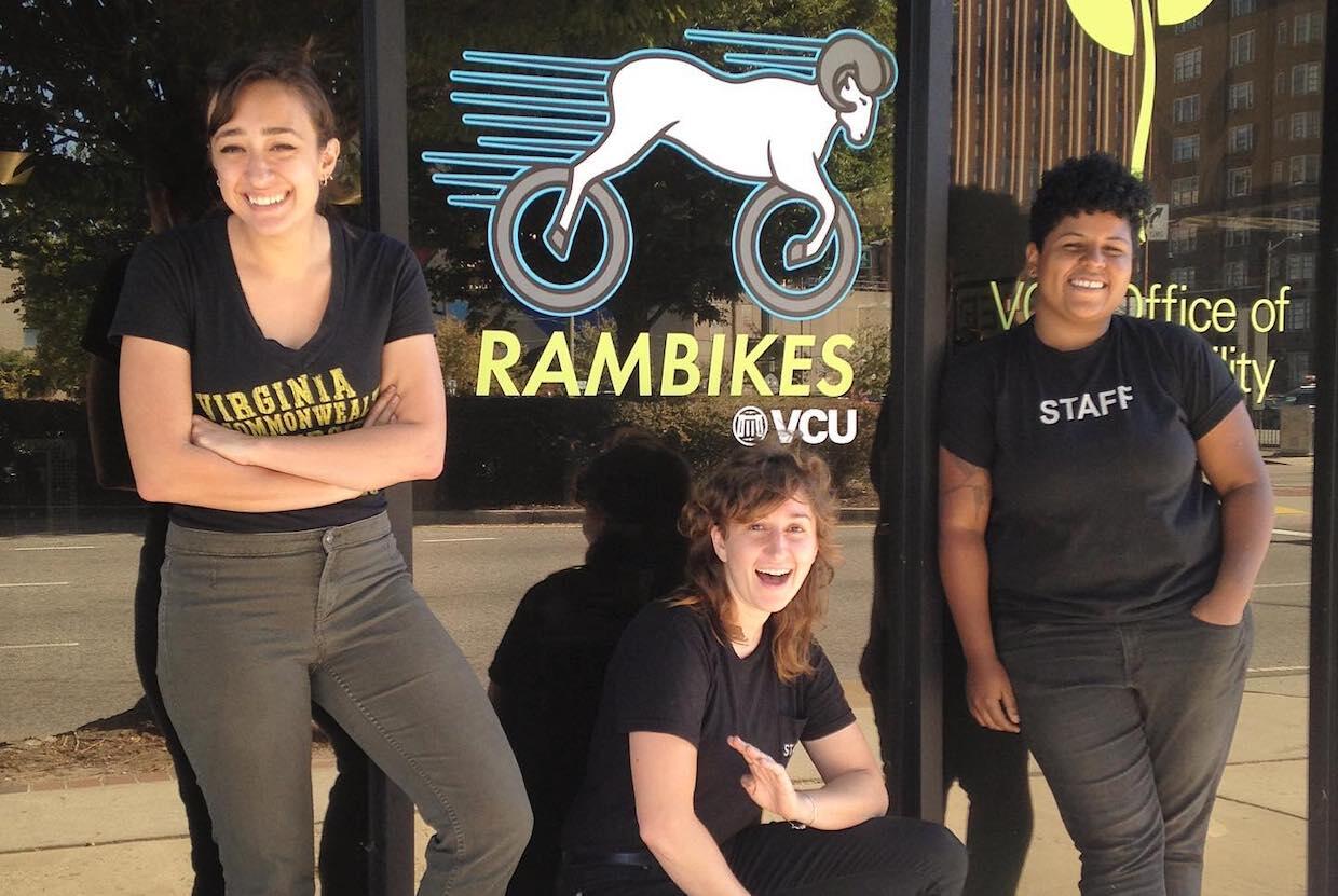 Three people stand outside of a store. The window displays the name and logo of the "Rambikes" shop at V C U.
