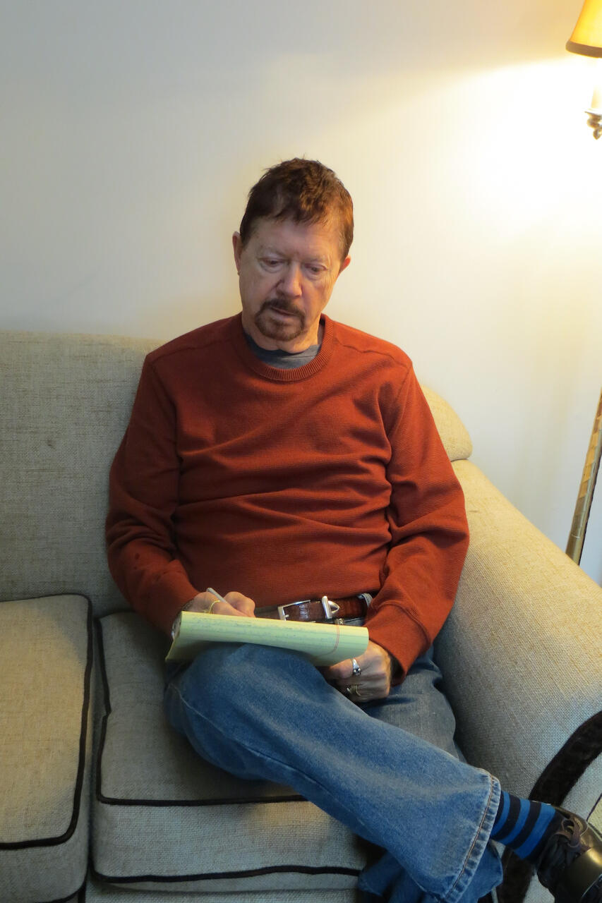 A photo of a man sitting on a couch and writing on a note pad. 