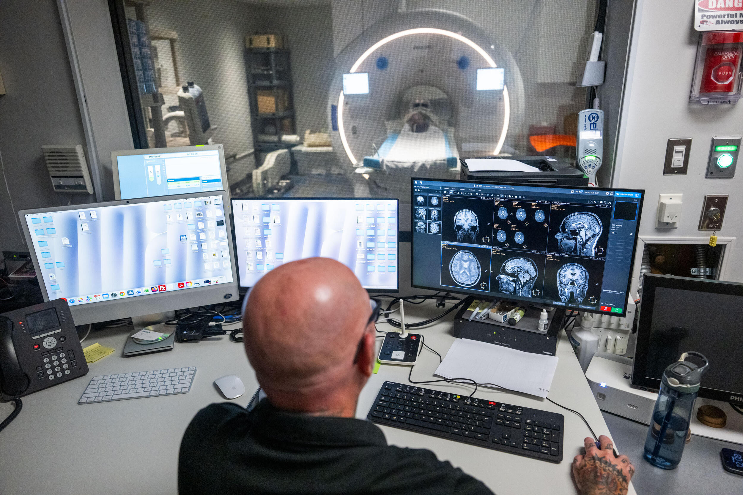 A photo of a man sitting at a desk with four computer monitors on it. Behind the desk is a galss window that shows and MRI machine on the other side.