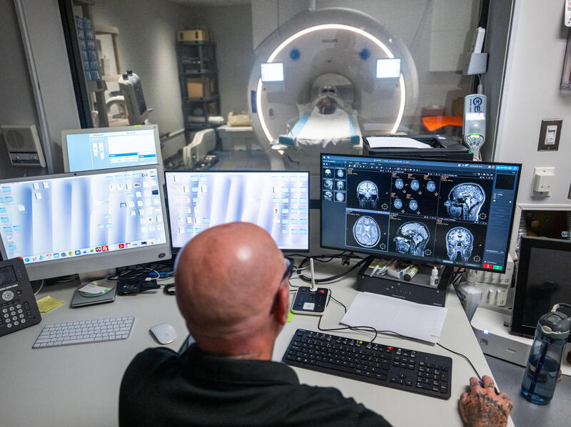 A photo of a man sitting at a desk with four computer monitors on it. Behind the desk is a galss window that shows and MRI machine on the other side. 