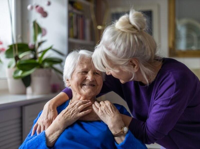 A photo of a woman wrapping her arms around an elderly woman with gray hair. Both women are smiling. 
