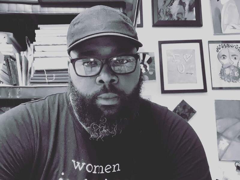 Amiri Richardson-Keys sitting in front of a bookcase and pictures hanging on a wall. His shirt says \"women inspire nations.\" 