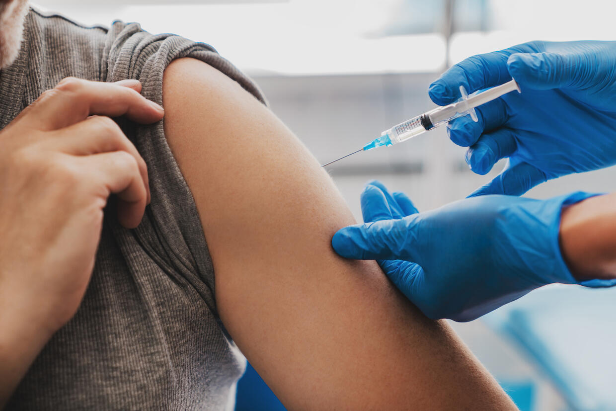 A photo of a person lifting up the sleeve of their shirt to reveal their right arm. Another person wearing blue latex gloves is giving a shot to the person with their arm exposed. 