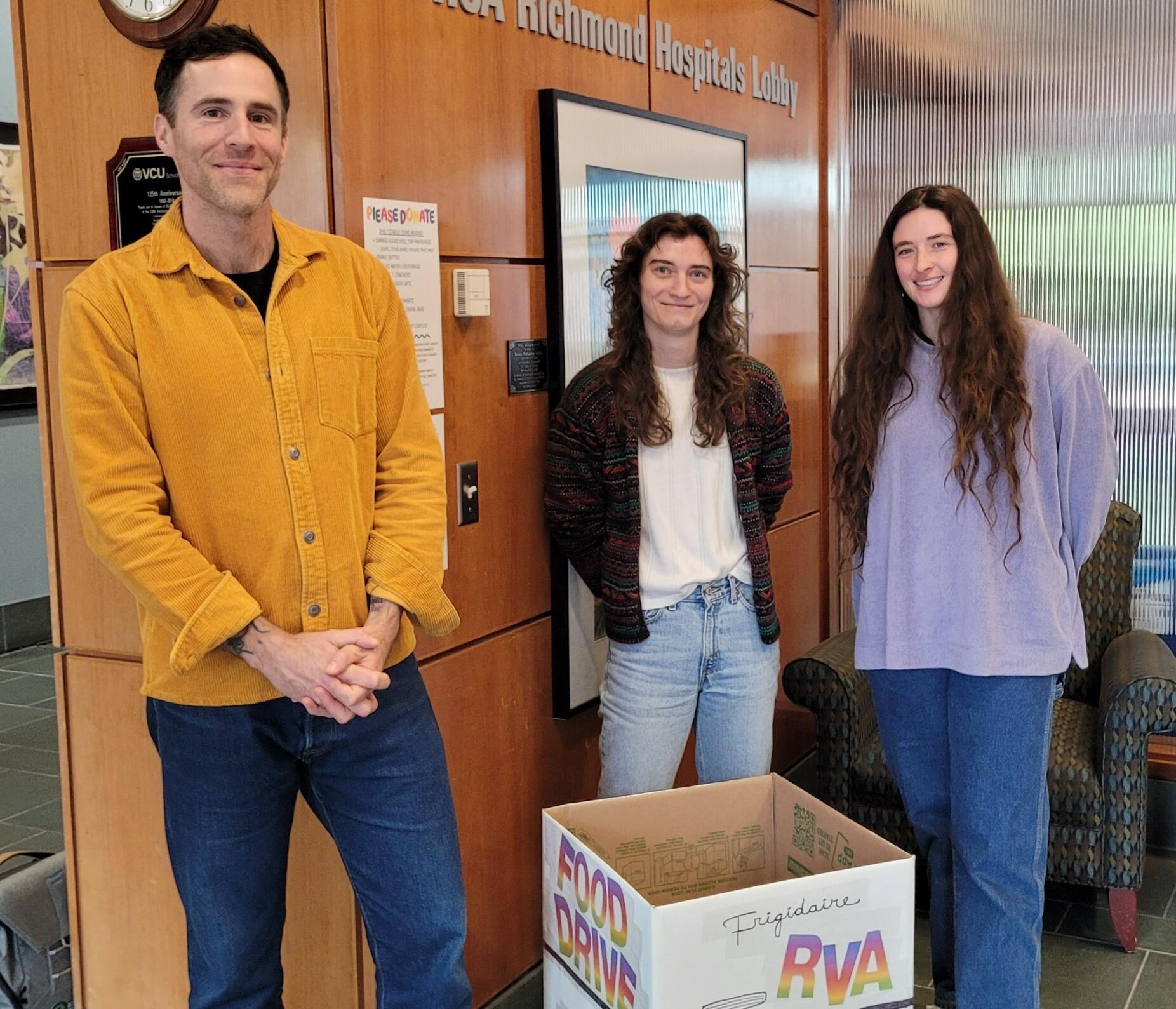 A photo of three people standing. On the left is a man and in the middle and right are two women. There is a white box in front of the group which says \"FOOD DRIVE\" and \"RVA\" in rainbow letters. 