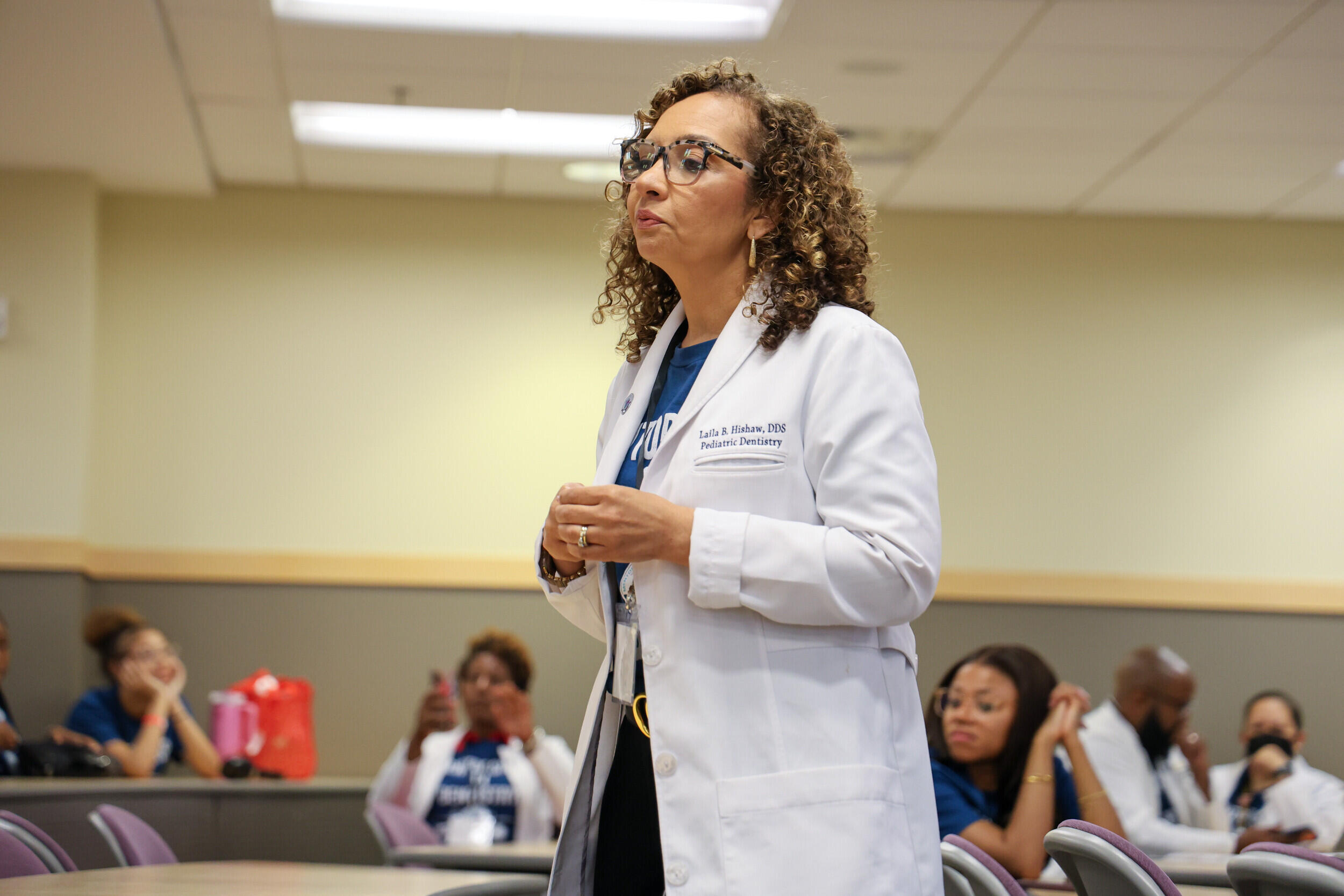 A photo of a woman standing in the middle of a lecture hall. There are people sitting at tables behind her. 