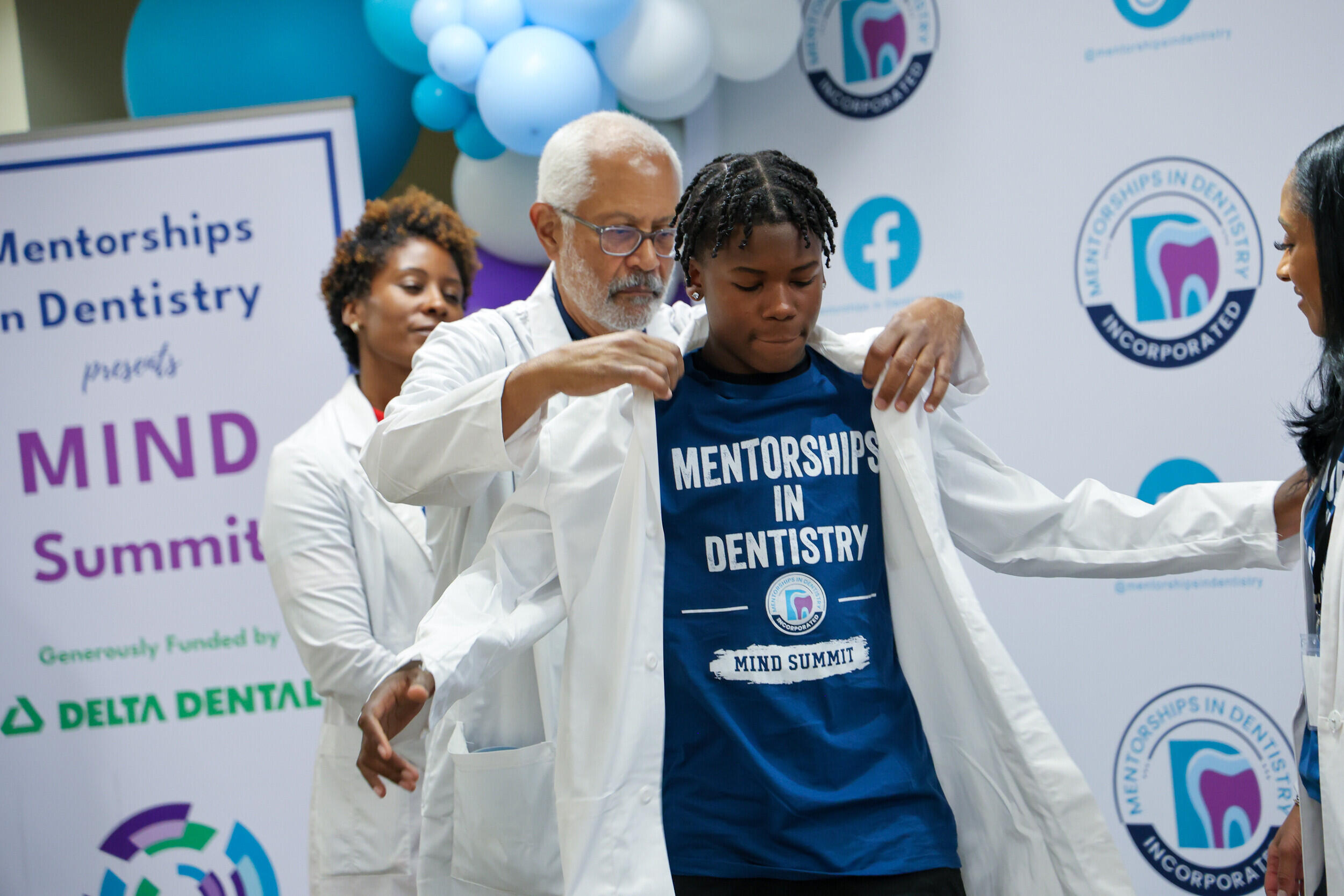 A photo of a man putting a white dentist coat on a younger man. There are two women in white dentist coats watching. 
