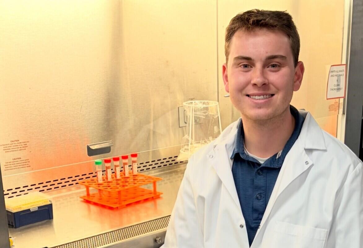 A phto of a man wearing a lab coat sitting in front of an open air vent with test tubes in it. 