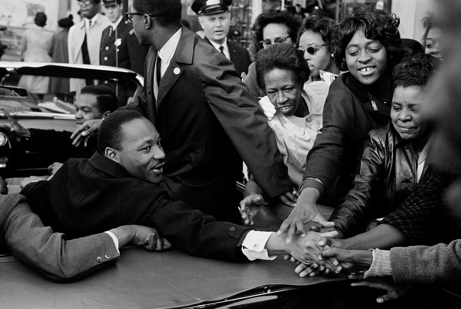 Martin Luther King Jr. reaches from a convertible toward onlookers with outstretched hands.