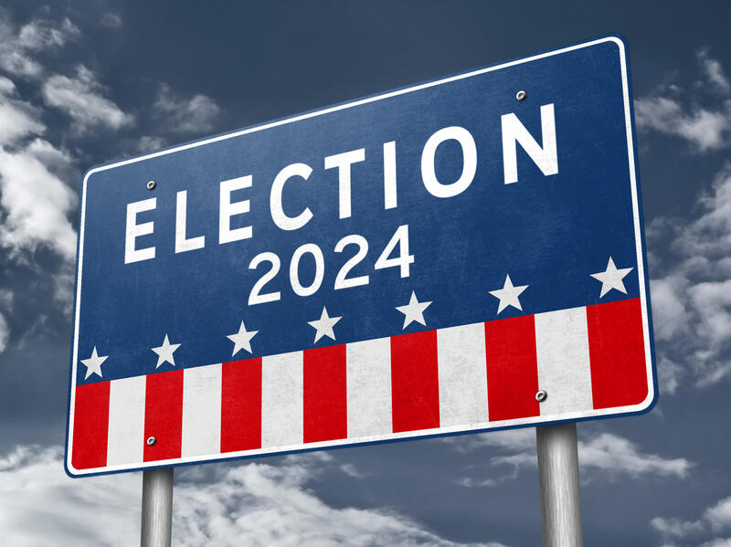 A photo of a road sign that says \"ELECTION 2024\" in white letters. The top of the sign is blue, with a row of white stars under the letters. Under the blue, are white and red stripes, making the sign imitate an American flag. 