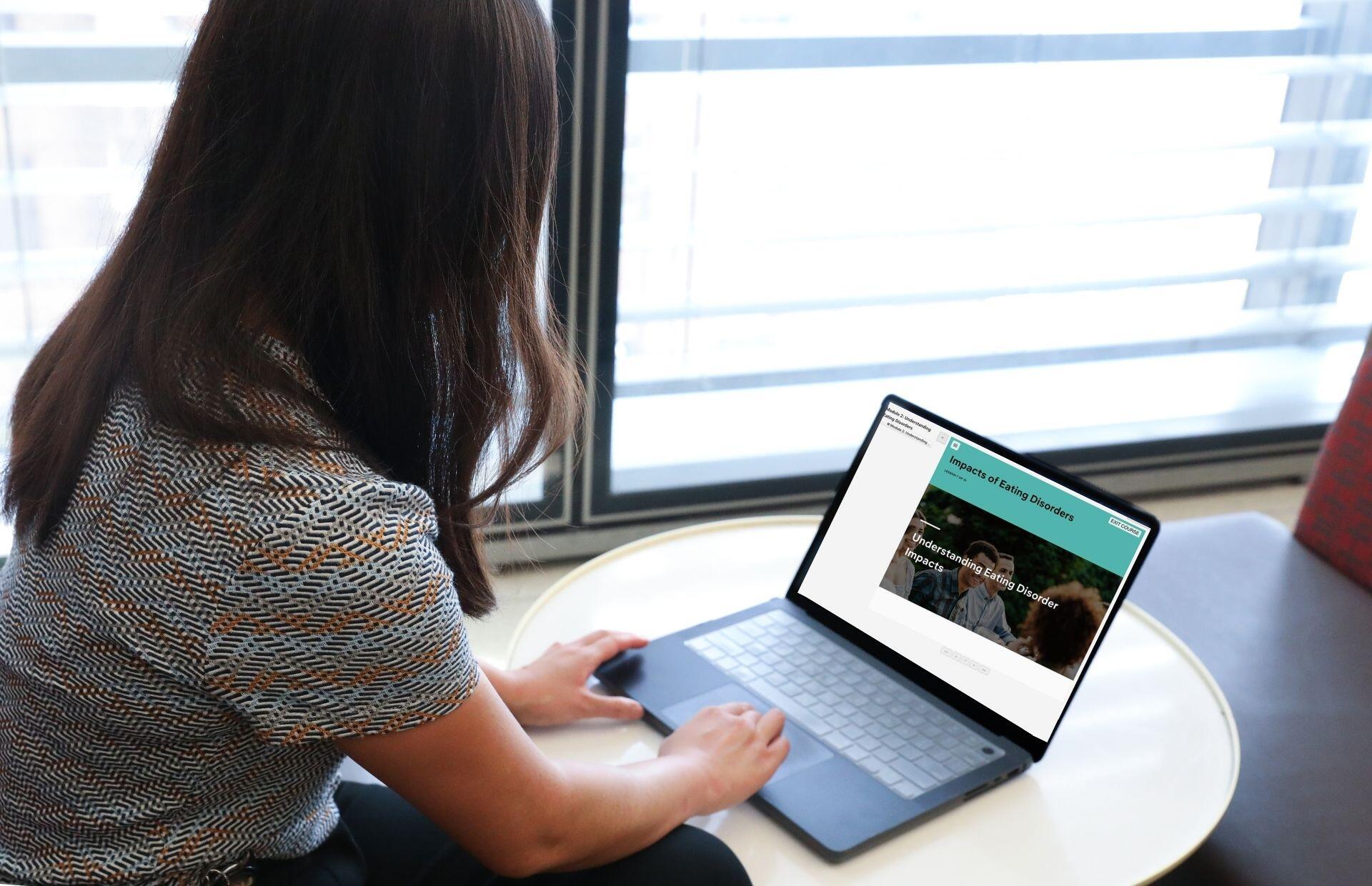 A photo of a woman from behind. She is sitting at a small table with a laptop on top of it. 