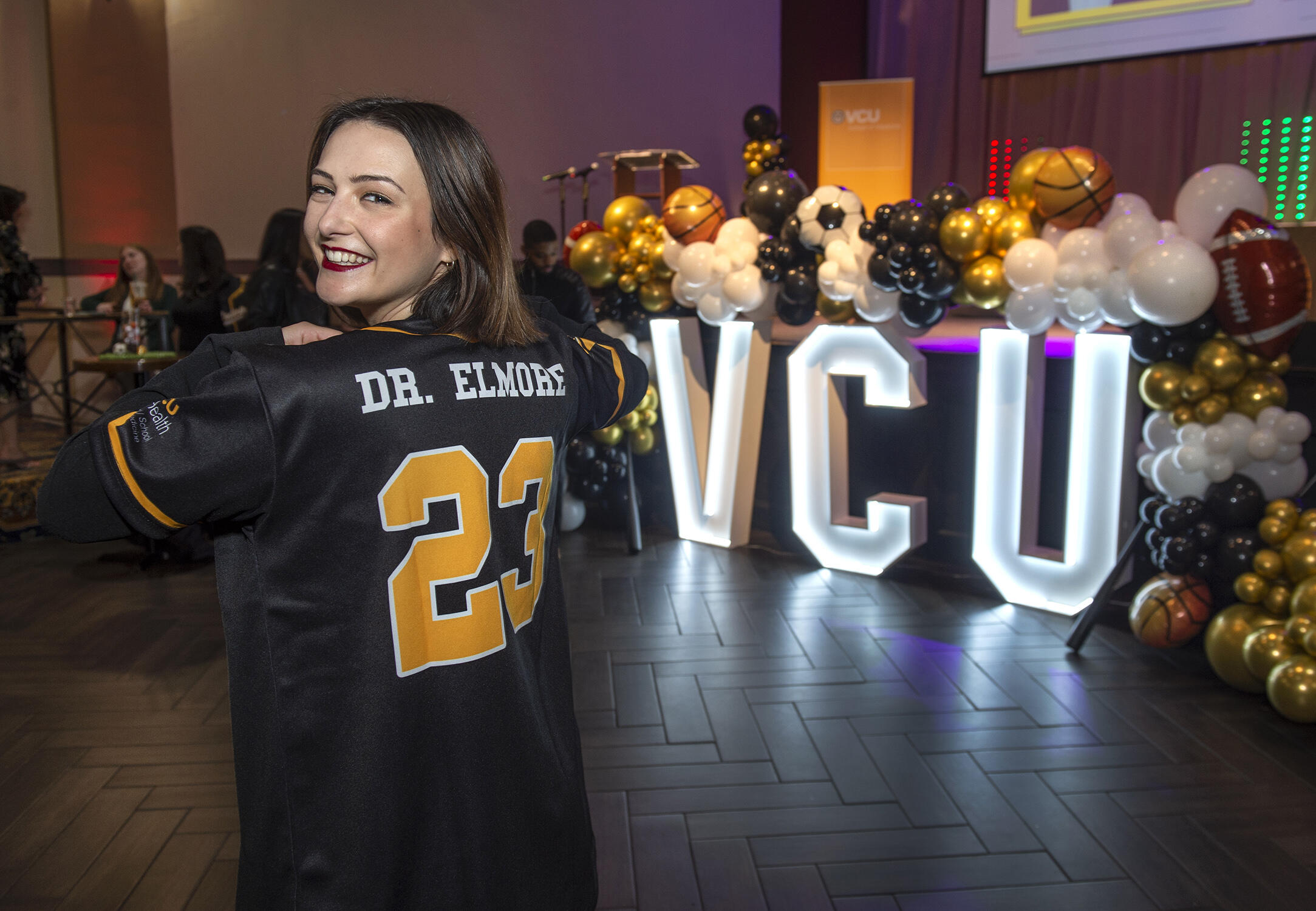 A woman looking behind her wearing a sports jersey that says \"DR. ELMORE 23\". To the woman's right are large white light up letters that spell out \"V C U.\" 