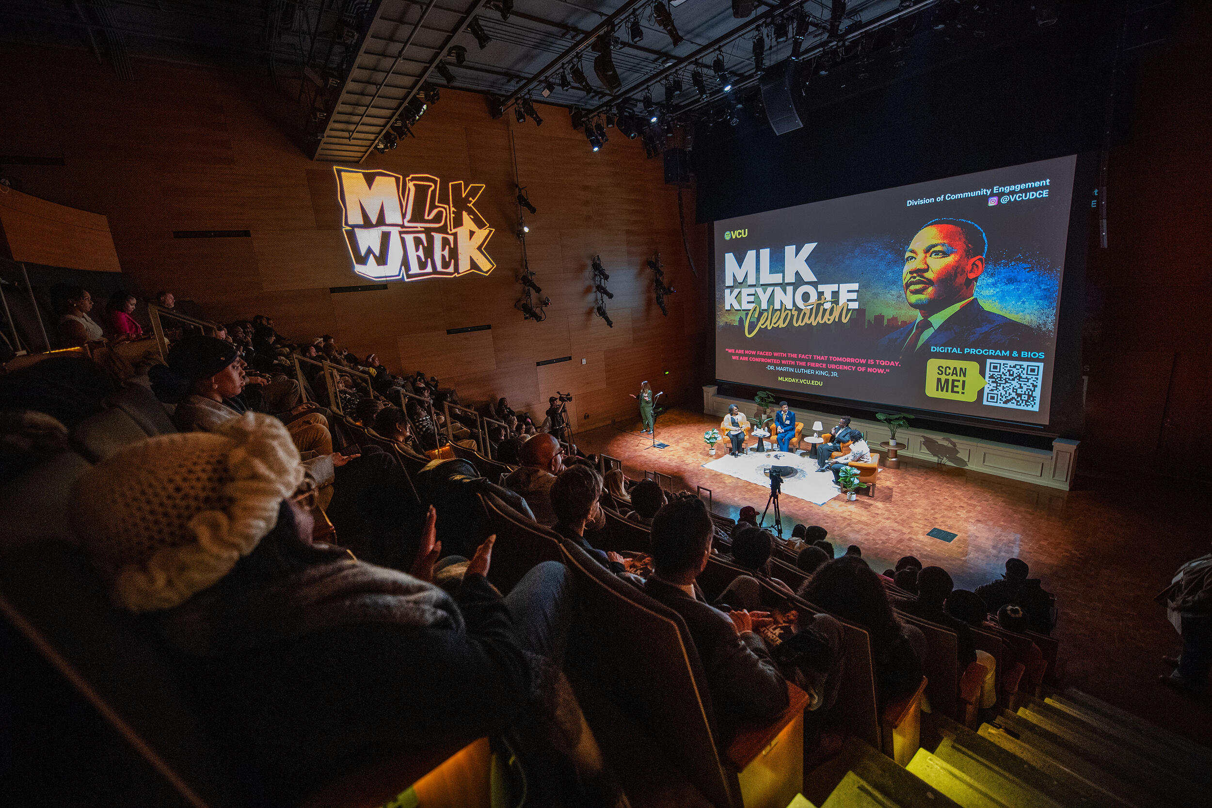 A photo of an auditorium filled with people sitting. At the front of the room are four people sitting in plush chairs in a semi-circle. 