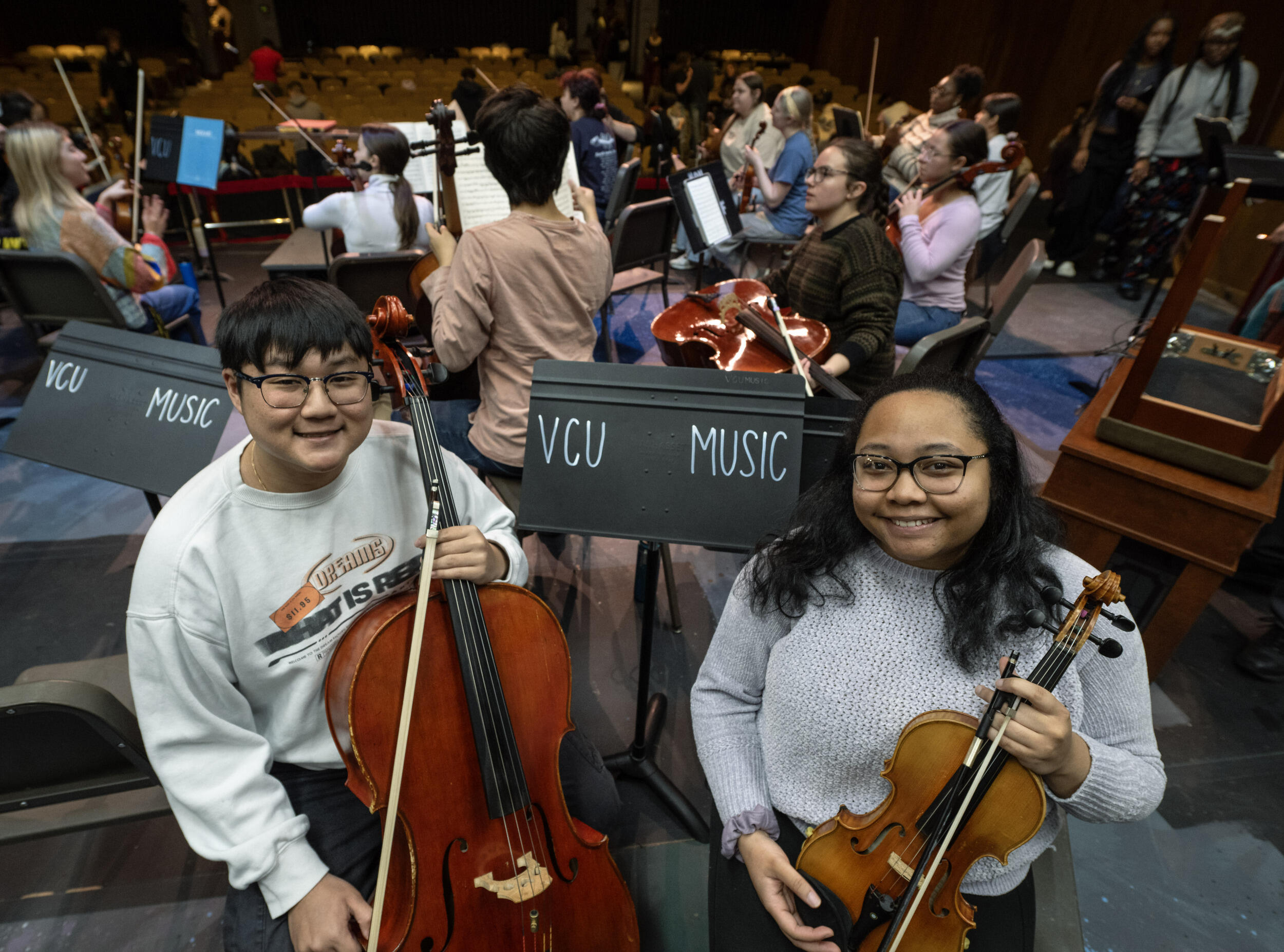 A photo of a man and a woman from the waist up holding string instruments. Behind them are two music stands that say \"VCU MUSIC\" in white letters. Behind the stands are a group of people standing holding musical instruments. 