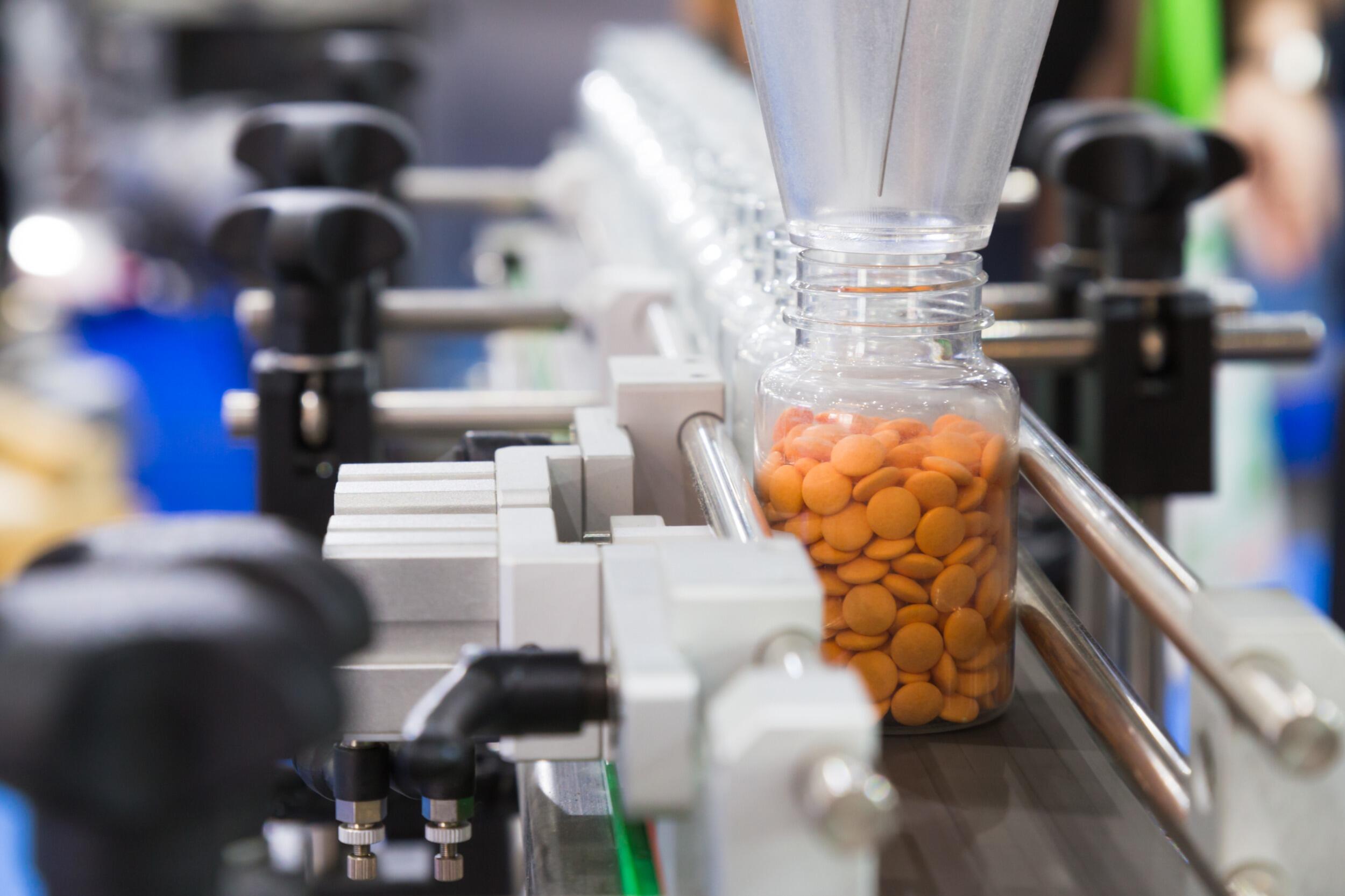 A manufacturing facility produces pharmaceutical products, including a bottle of orange pills, on a machine.
