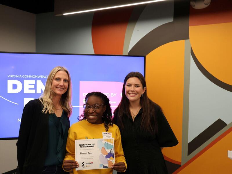 Three women smile in a row. The one in the middle is holding up a certificate.