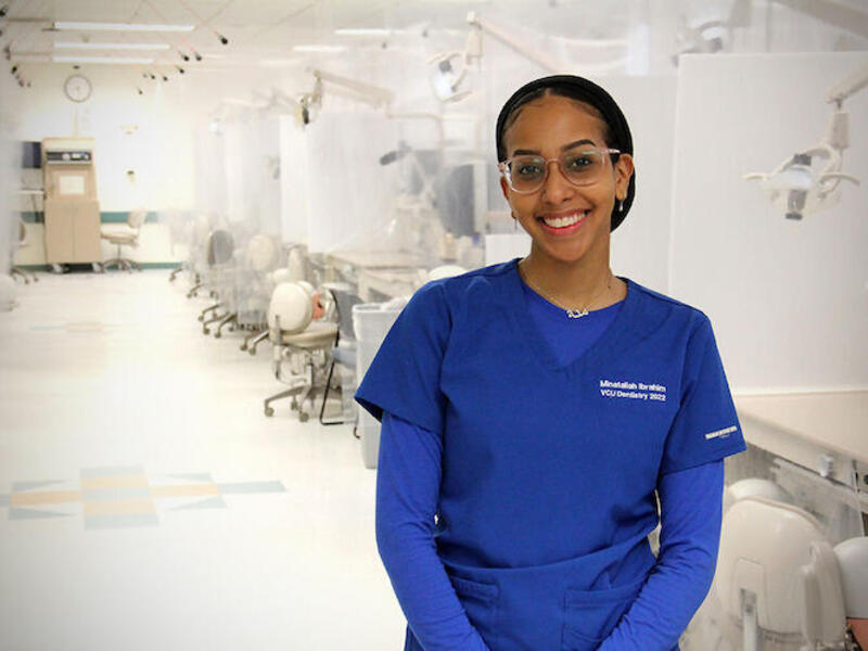 A woman in scrubs and glasses standing in front of a row of dental exam spaces. 