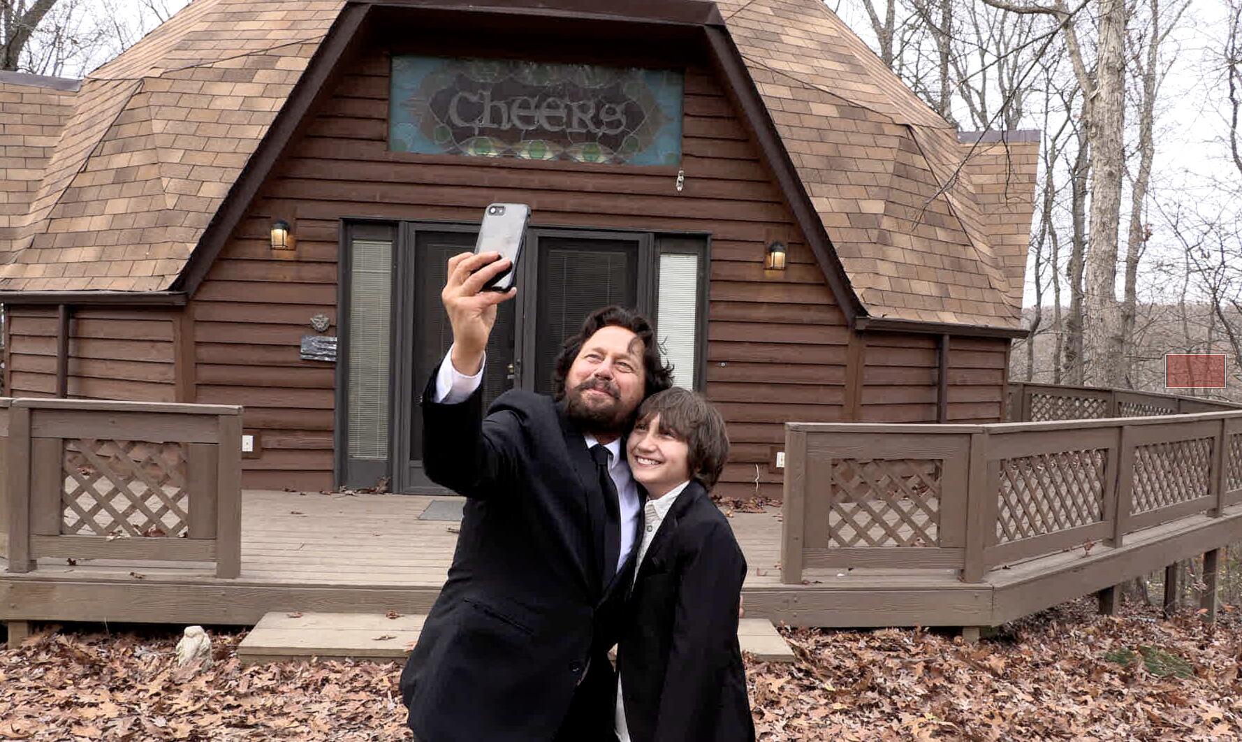 a man and a young boy take a selfie in front of a cabin