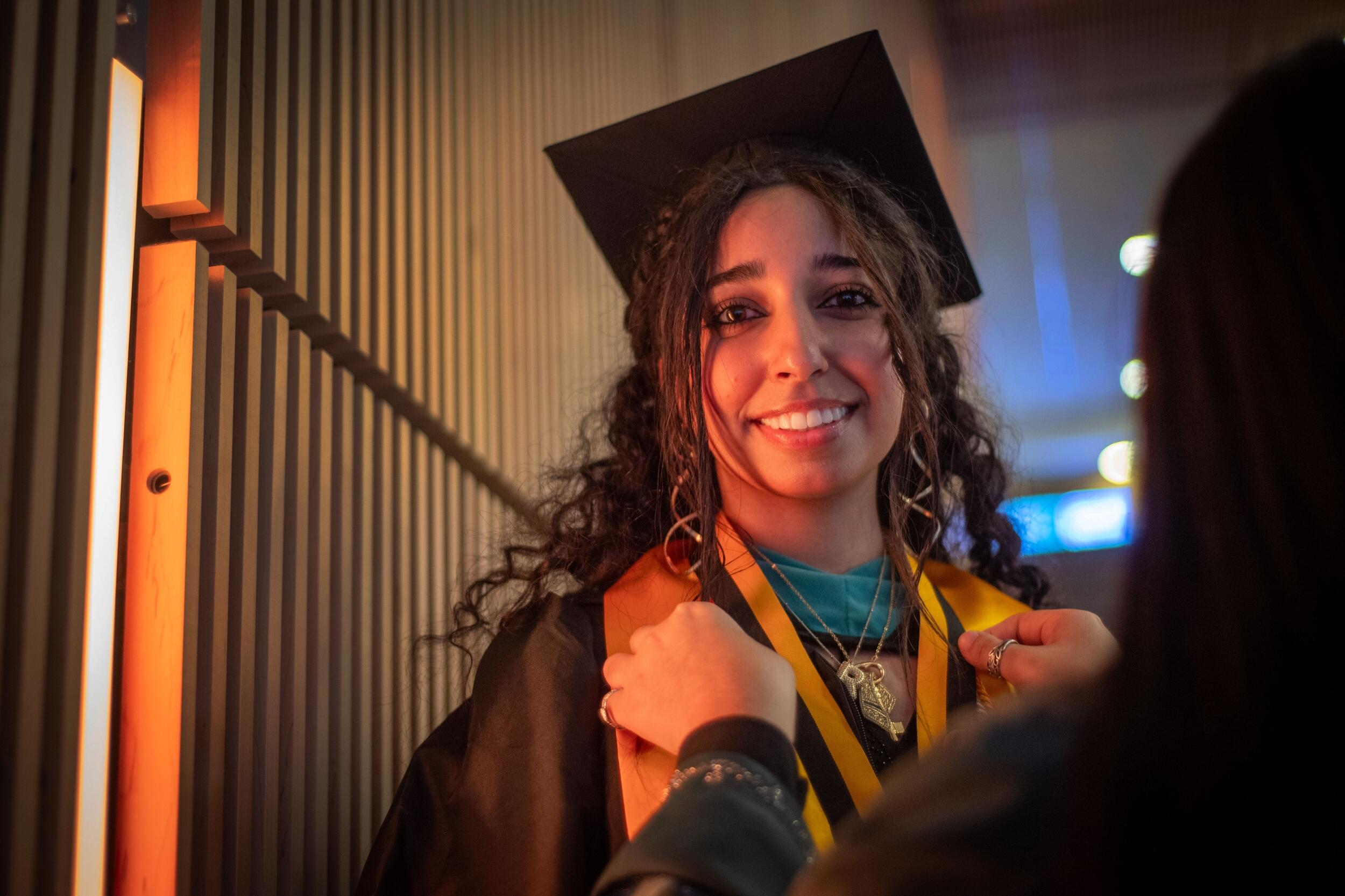 A photo of a woman wearing a graduation cap and gown having another person put a ribbon around her neck. 