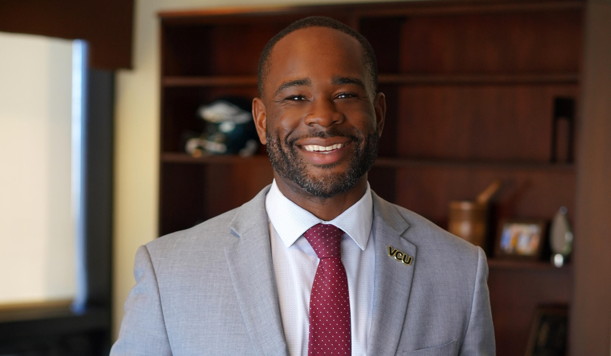 A portrait of a smiling man wearing a light gray sports jacket, white button up shirt, and burgundy tie. On his lapel is a pin that says \"VCU\". 