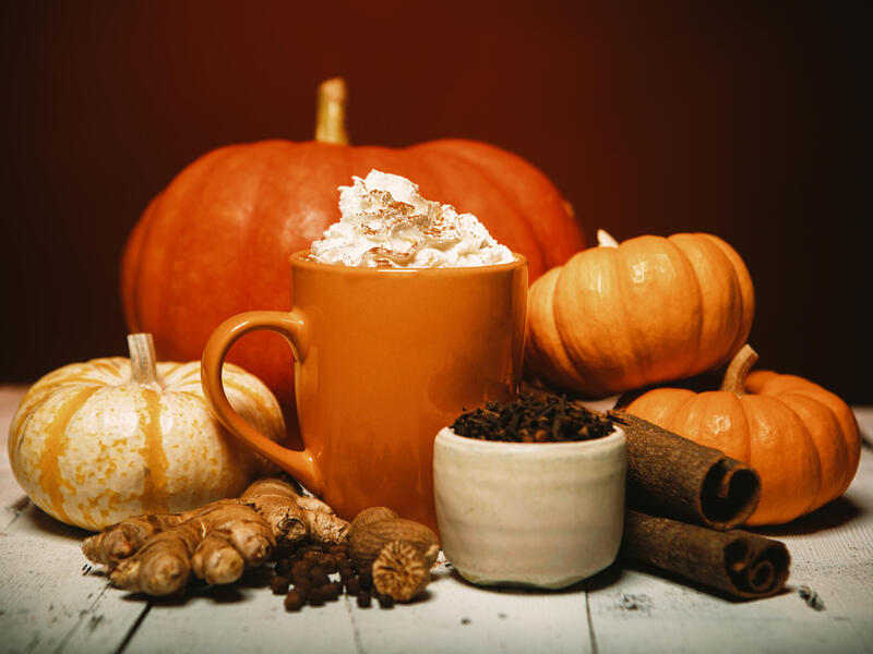 A photo of an orange mug topped with whipped cream surrounded by spices like cinnamon, all spice, and ginger. Behind the cup are four pumpkins. 