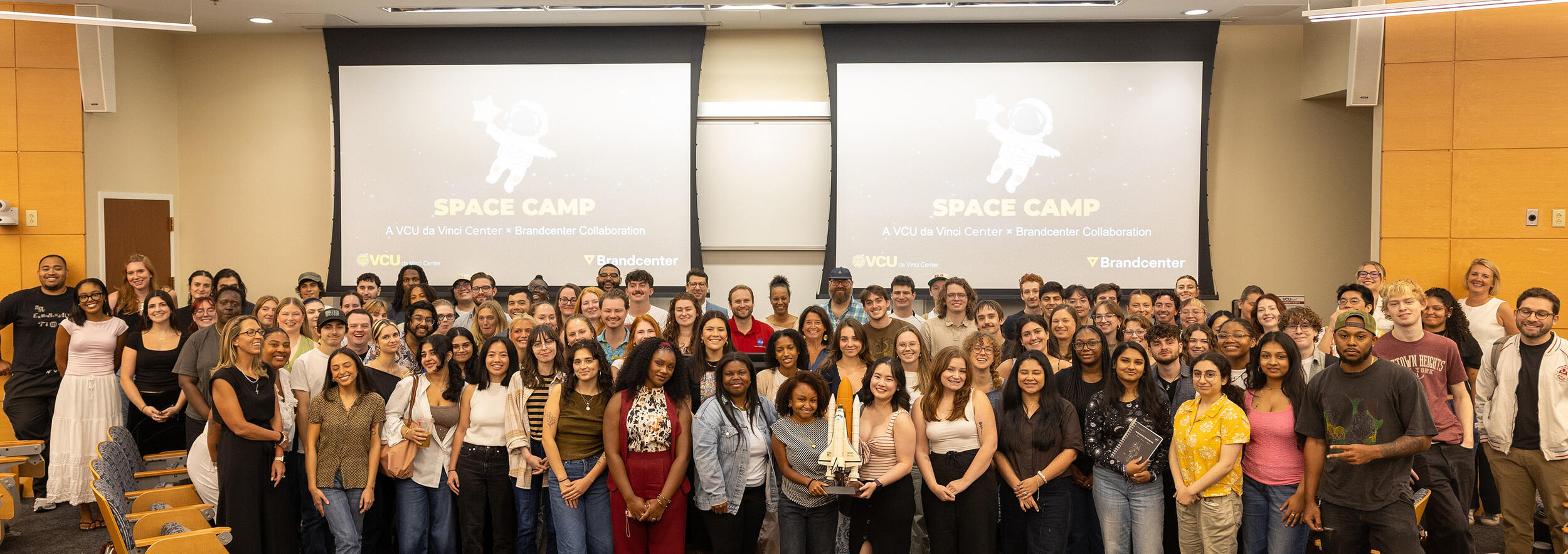 A group photo of dozens of people standing at the front of a room. Behind the people are two screens that say \"SPACE CAMP\" in yellow letters with an illustration of an astronaut above them. 