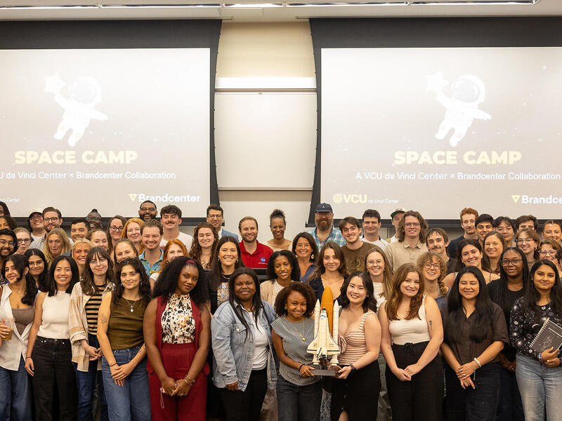 A group photo of dozens of people standing at the front of a room. Behind the people are two screens that say \"SPACE CAMP\" in yellow letters with an illustration of an astronaut above them. 