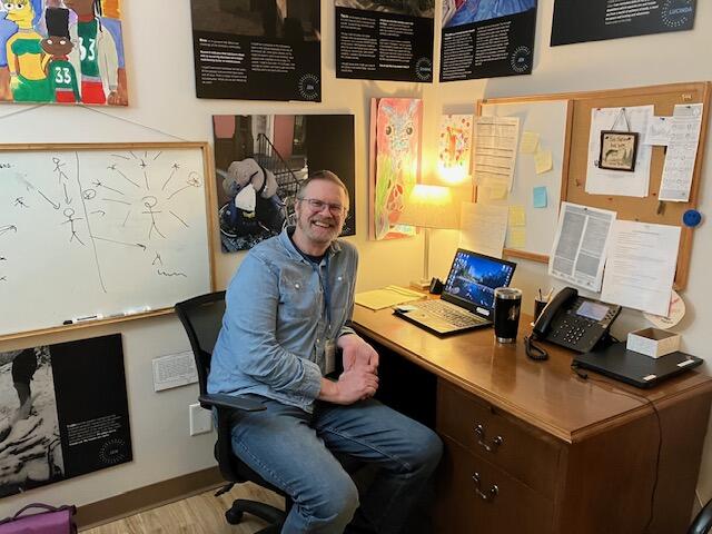 A photo of a man sitting at a desk and smiling. 