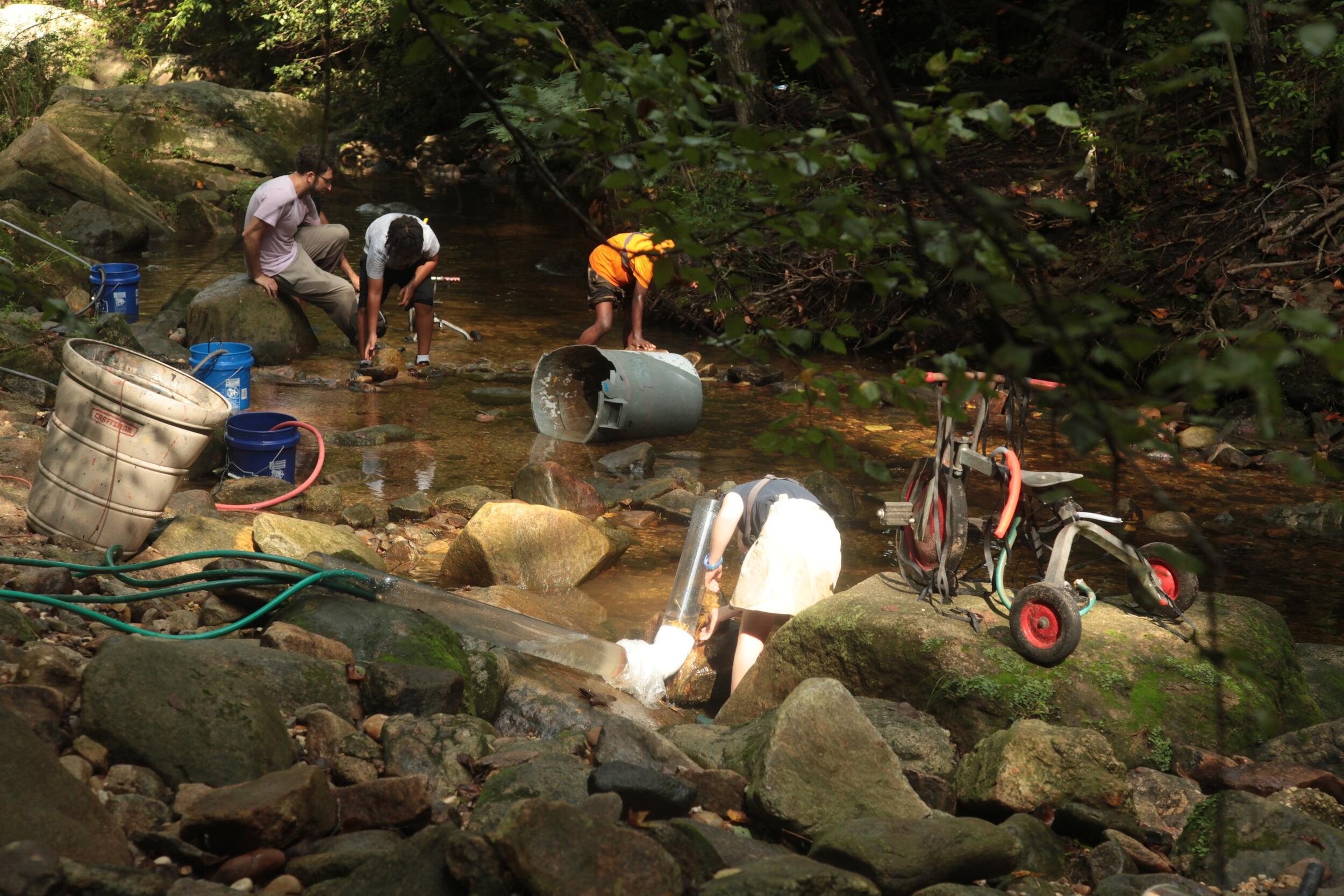 A photo of children playing in a stream. There is a bike sitting on a rock, a trashcan in the stream, as well as hoses and pipes. 