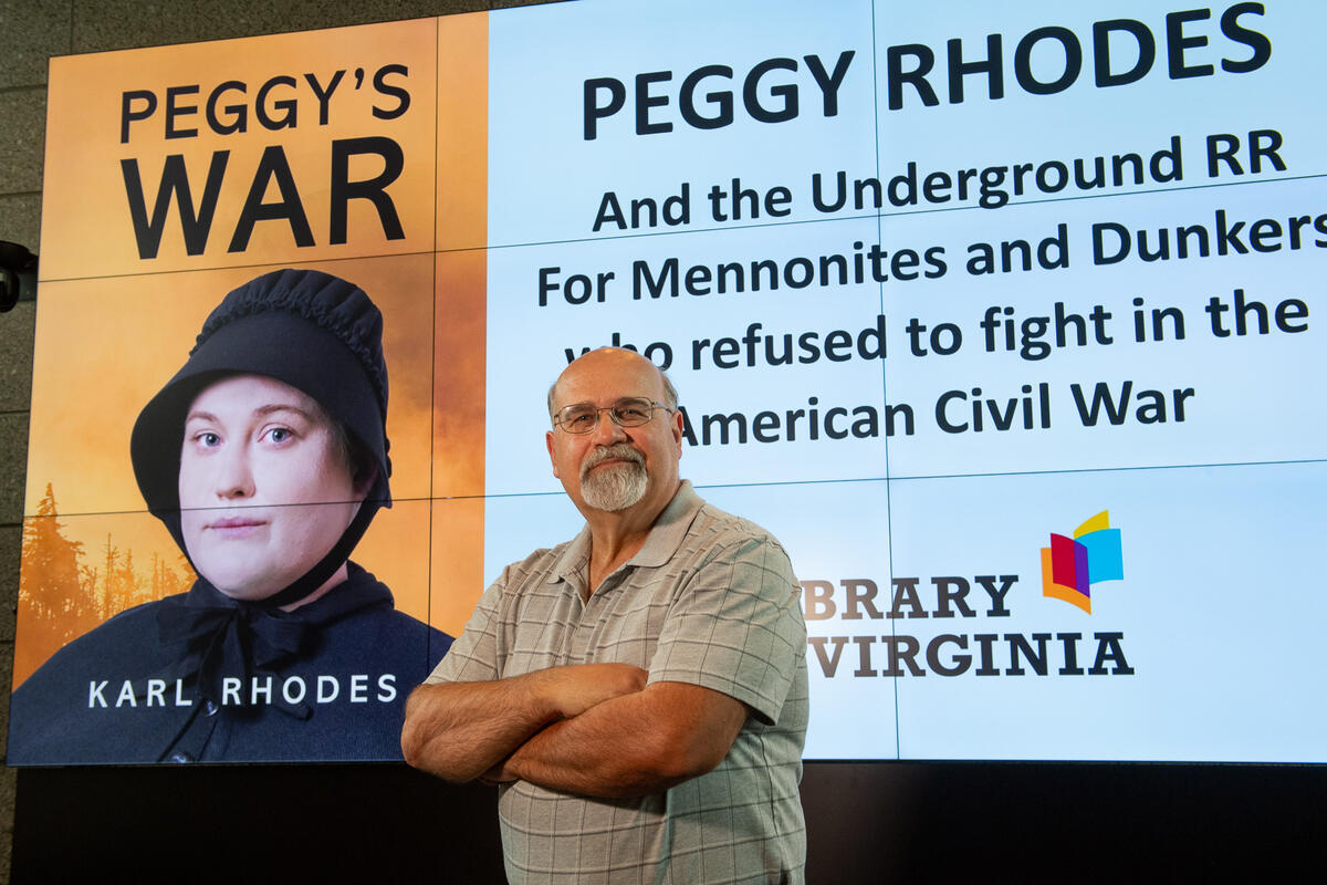 A photo of a man from the waist up standing with his arms crossed against his chest and smirking. Behind him is a screen that says \"PEGGY RHODS And the Underground RR For Mennonities and Dunkers who refused to fight in the American Civil War\" in black text with a photo of the book cover for \"Peggy's War\" next to it. 