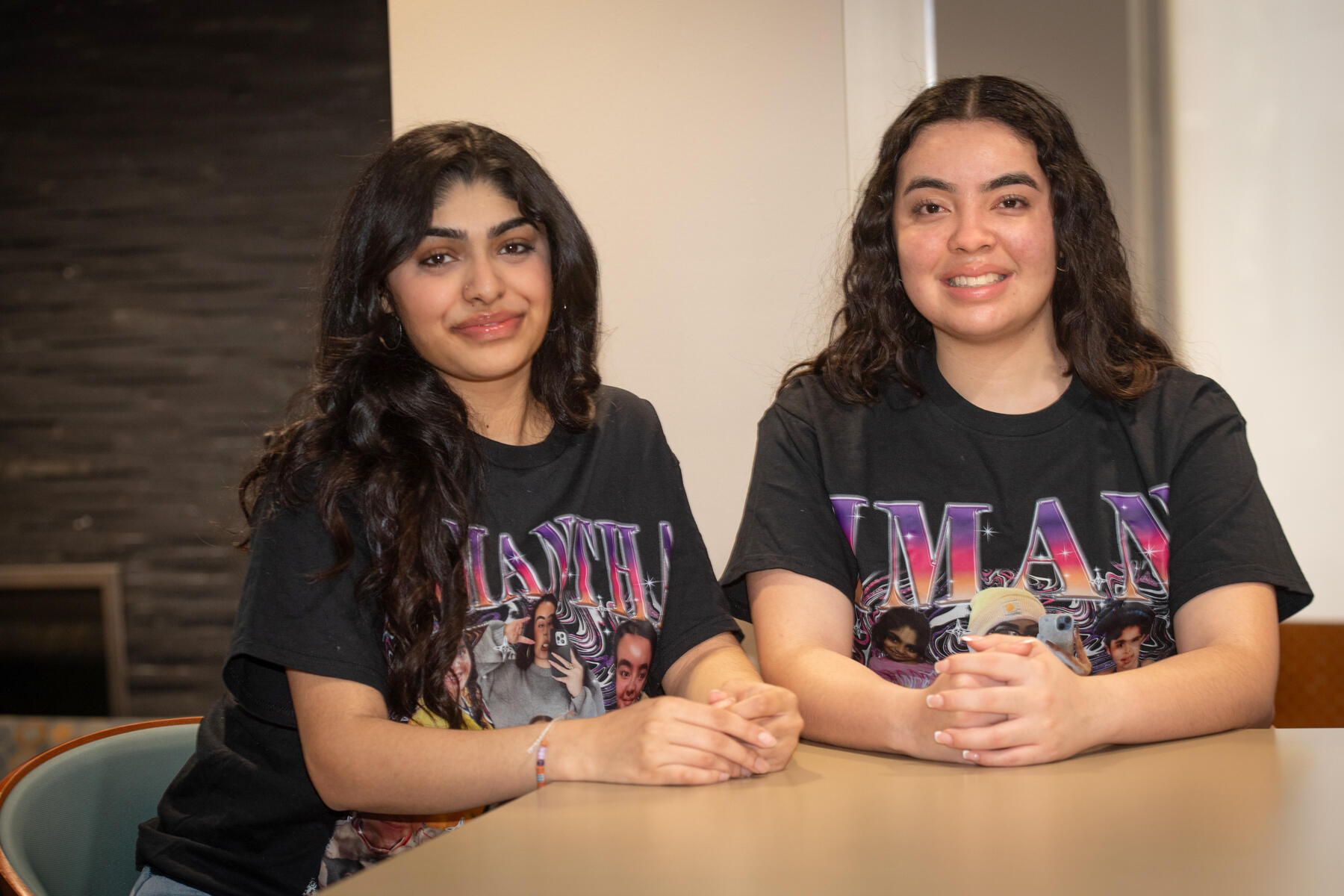 Two women sit at a table. Both wear black T-shirts -- one with the name Samantha on it, and the other with the name Iman on it.