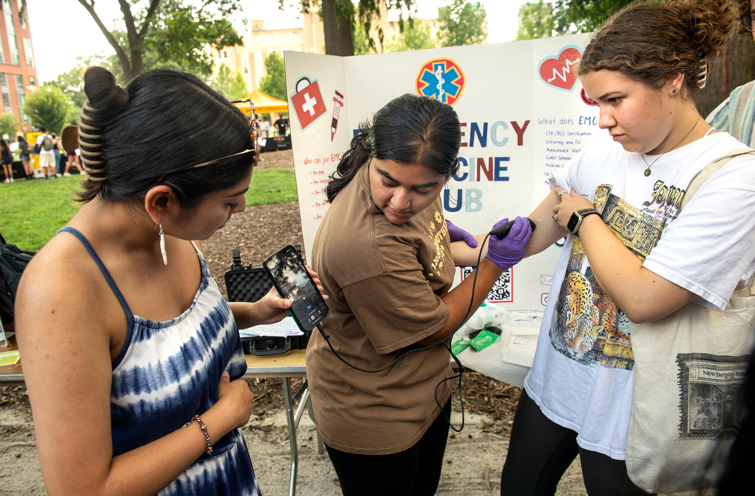A student watches as one student demonstrates how to use a handheld device by practicing on the inside of another student's arm to observe the student's vital signs. All three stand in front of a poster that says Emergency Medicine Club on a sidewalk next to a tree-lined green space in Monroe Park.