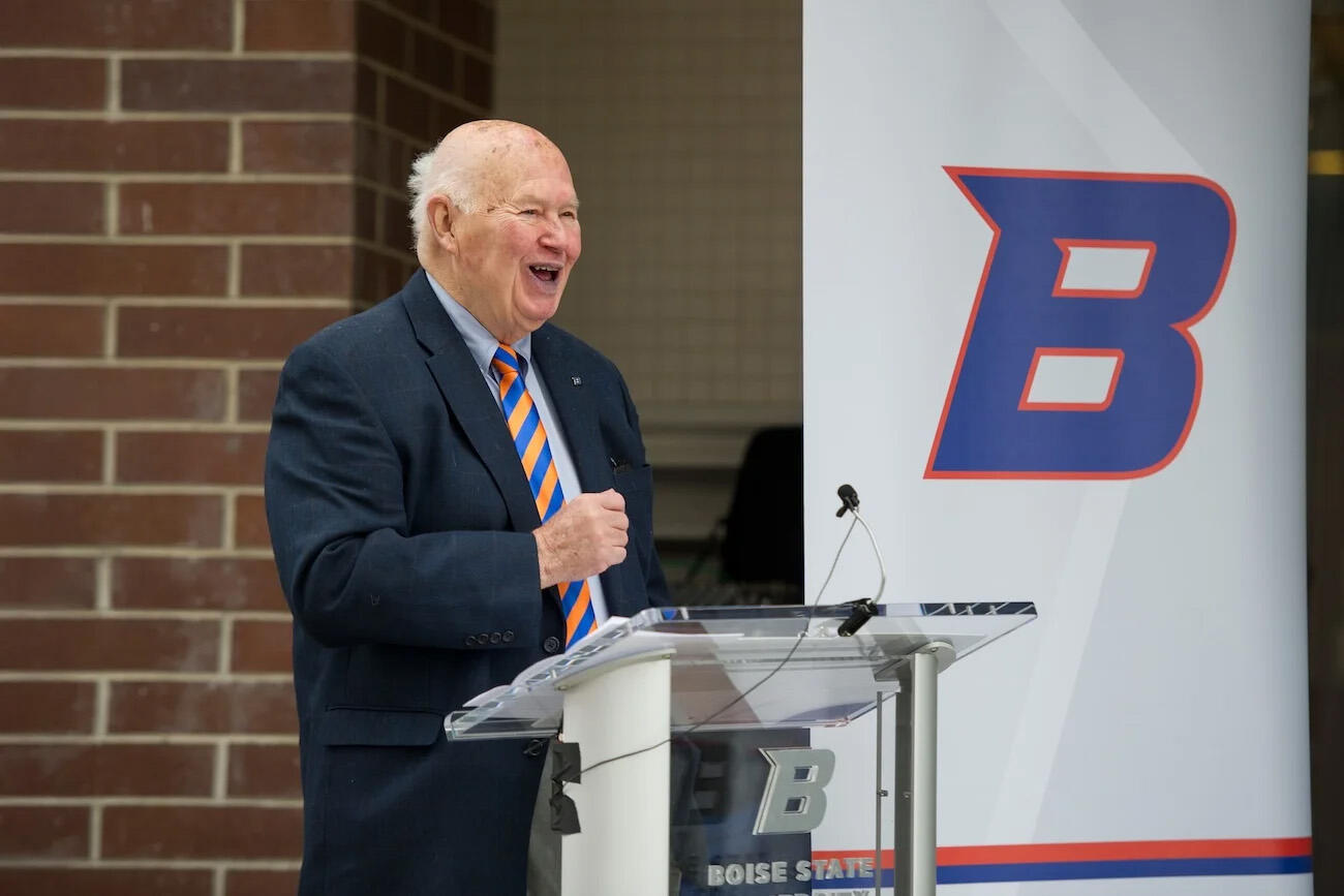 A photo of a man from the waist up. He is standing behind a podium with a microphone on it. Next to him is a white banner with the letter \"B\" on it. The letter is blue with a think orange outline. 